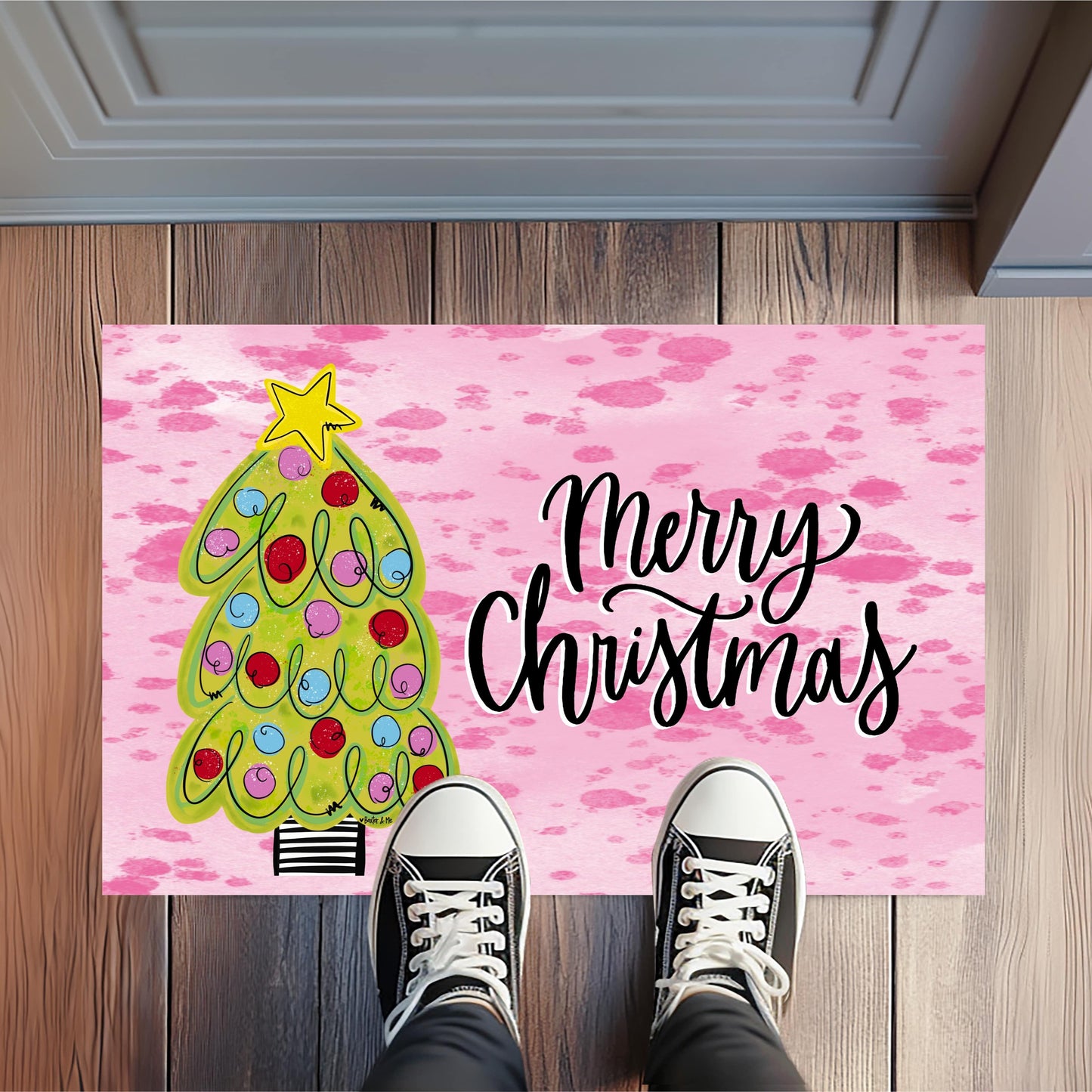 Pink doormat with Christmas tree and 'Merry Christmas' text on a wooden floor.