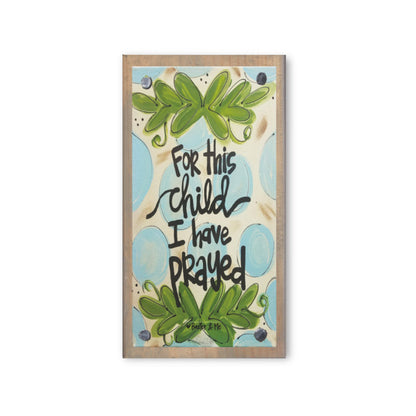 A wooden block with a printed design featuring leaves and the text 'For this child I have prayed' in a cursive and decorative font, with a blue background.