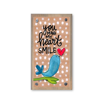 A wooden block with a blue bird and a heart, and the text 'you make my heart smile' printed on it, against a beige background with white polka dots.