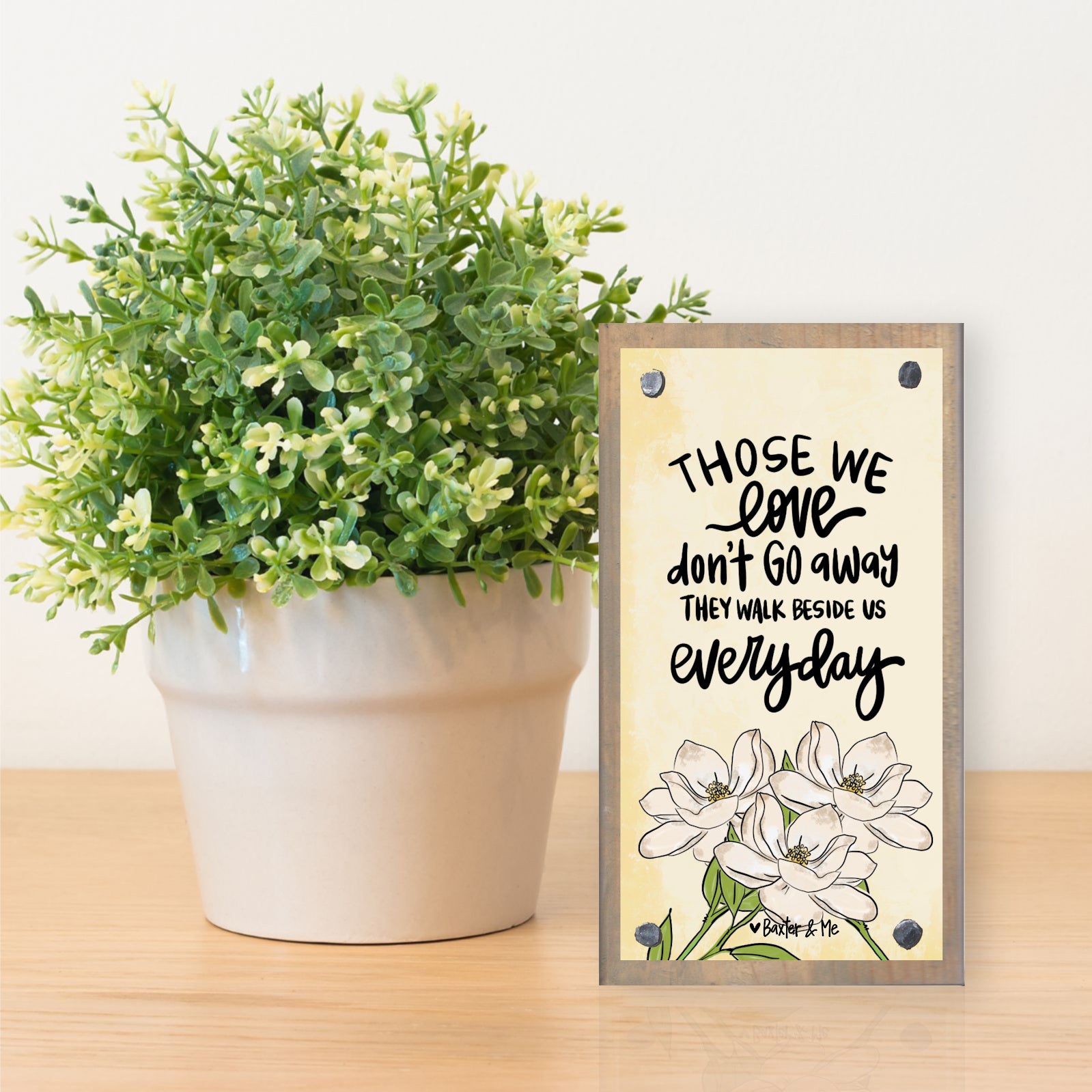 Decorative sign with floral design and text next to a potted plant on a wooden surface.