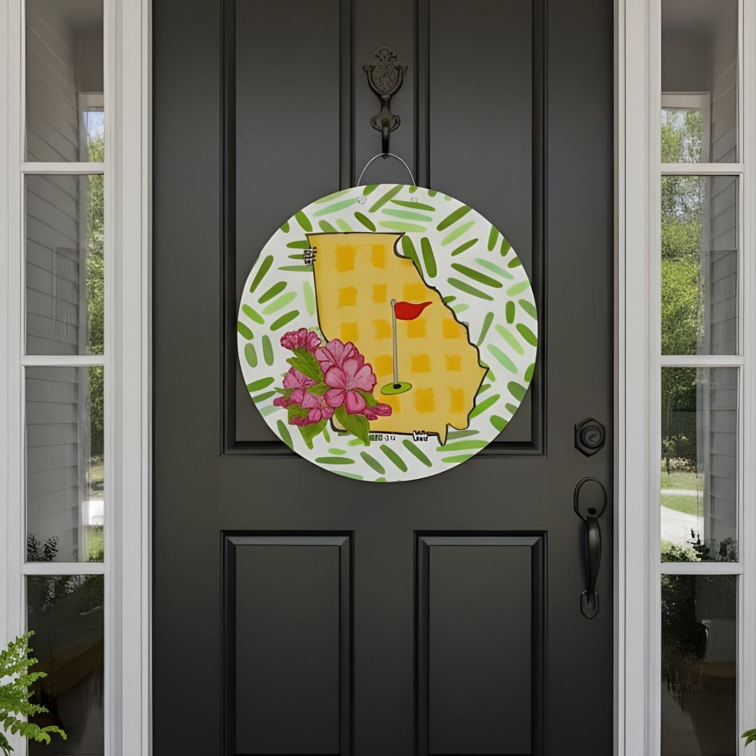 Decorative door hanger with a yellow Georgia state outline, pink flowers, a green golf flag and green leaves on a black door.
